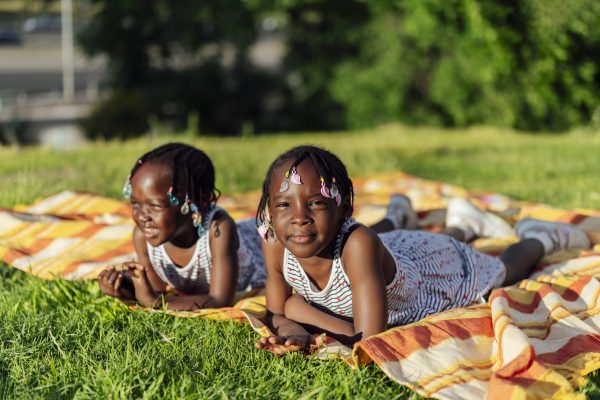 Two african twin sisters relaxing on picnic blanket in park during summer day